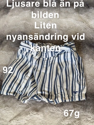 Randiga shorts med knytband - Säljer ett par vita och blå randiga shorts med knytband i midjan. Shortsen har en loose passform och elastisk midja för extra komfort. Perfekta för varma dagar och har en avslappnad stil.🌸 Liten nyansändring vid kanten. Ljusare blå än på bilden. Storlek 92