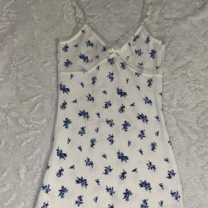 Vit blommig kortklänning från H&M Divided - Supersöt vit kortklänning med blått blommönster från H&M Divided. Klänningen har smala axelband, spetsdetaljer vid bysten och en liten rosett framtill. Skriv vid frågor 🥰