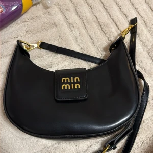 Svart axelväska - Snygg svart axelväska från Min Min i slät skinnimitation med guldfärgade detaljer och logga framtill. Väskan har en avtagbar axelrem och en stilren, rundad form som ger en modern vibe. Perfekt för dig som vill ha en clean och trendig accessoar.