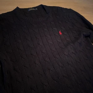 Mörkblå stickad tröja från Polo Ralph Lauren - Kabelstickad mörkblå tröja från Polo Ralph Lauren med klassisk röd logga broderad på bröstet. Tröjan har rund halsringning och långa ärmar, perfekt för lager på lager under kyliga dagar.