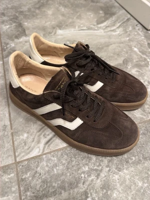 Gant Sneakers Cuzima - Säljer ett par bruna Cuzima sneakers från Gant i mocka med vita detaljer och gummisula. Skorna har snörning, rund tå och vita ränder på sidan samt vit häl. Perfekta för dig som gillar retrovibbar och stilren design. Använda 2 gånger max, säljes pga av att de inte passar min klädstil 🤎köpta för 1700 men kom med prisförslag 