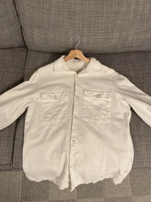 Vit overshirt med fickor - Säljer en stilren vit overshirt med två stora bröstfickor och knappar framtill. Skjortjackan har klassisk krage och är långärmad. Perfekt att slänga över en t-shirt för en clean look.