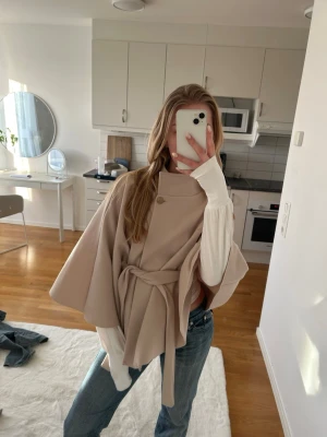 Beige kappa med bälte och guldknapp - Säljer en beige kappa med snyggt bälte i midjan och en dekorativ guldig knapp vid halsen. Kappan har en cape-liknande form med vida ärmar och är tillverkad i ett mjukt ull-liknande material. Perfekt för dig som vill ha en stilren och trendig look.