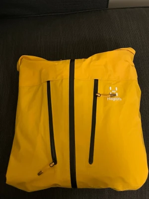 Gul regnjacka från Haglöfs dam L - Säljer en gul regnjacka från Haglöfs i dammodell, storlek L. Jackan har svarta dragkedjor framtill, hög krage och justerbar huva. Materialet är slitstark polyester och jackan har två stora bröstfickor med dragkedja. Perfekt för blöta dagar!