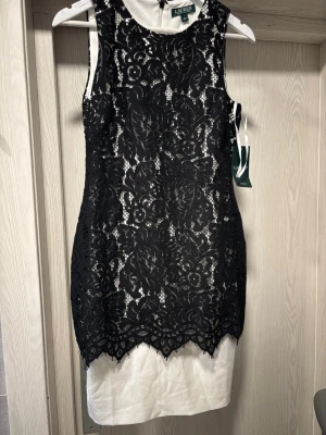 Svart spetsklänning från Lauren Ralph Lauren - Säljer en elegant kort klänning från Lauren Ralph Lauren med svart blommig spets över ett vitt foder. Klänningen är ärmlös, har rund halsringning och dragkedja i sidan. Perfekt för dig som vill ha en klassisk och stilren look.