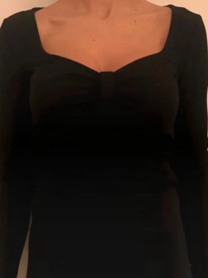 Svart långärmad topp med fyrkantig ringning - Svart långärmad topp med snygg fyrkantig ringning och en liten detalj framtill vid bysten. Toppens passform är figurnära och den är perfekt att styla med jeans eller kjol för en trendig look.