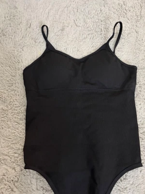 Svart ribbad bodysuit med smala axelband - Ser väldigt liten ut men meningen är att den ska sitta tajt! Har insydda bh-”kuddar”