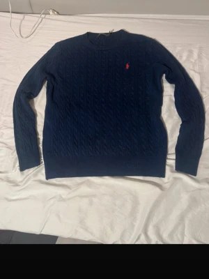 Mörkblå kabelstickad tröja Polo Ralph Lauren - Kabelstickad mörkblå tröja från Polo Ralph Lauren med klassisk rund hals och röd broderad logga på bröstet. Tröjan är långärmad och har ribbade muddar. Perfekt för dig som gillar stilren och tidlös design.