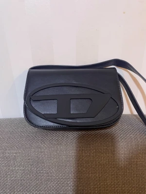 Svart axelväska från Diesel i skinn - Säljer en svart axelväska från Diesel i slätt skinn med ett stort, coolt Diesel-emblem på framsidan. Väskan har magnetlås, justerbar axelrem och ett ljust innerfoder. Perfekt storlek för det viktigaste och en stilren look till alla outfits.