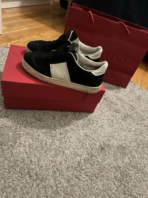 Valentino Garavani svartvita sneakers - Säljer ett par Valentino Garavani sneakers i svart mocka och vitt skinn med coola detaljer på sulan och metallnitar på sidan. Klassisk låg modell med snörning och rund tå. Snygg kontrast mellan svart mocka och vita partier. Kommer med originalkartong och påse. Väldigt prisvärt.