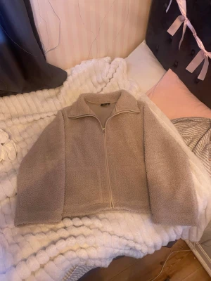 Beige teddyjacka från lager 157, storlek M - Mysig beige teddyjacka från lager 157 i storlek M. Jackan har dragkedja framtill, hög krage och två fickor. Perfekt för kyliga dagar när du vill vara både varm och snygg. Passar till allt och är superenkel att styla.