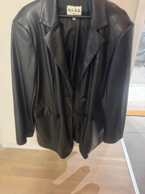 Svart skinnkavaj från NA-KD - Säljer en svart kavaj i skinnimitation från NA-KD. Jackan har klassisk krage, knäppning framtill med två knappar och fickor på sidorna. Långärmad modell med clean look och snygg passform. Storlek 38 Oversized i modellen.