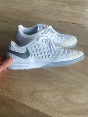 Vita Nike Lunar Gato II fotbollsskor - Säljer ett par vita Nike Lunar Gato II inomhusfotbollsskor med silverfärgad swoosh. Skorna har quiltad tå, meshpaneler på sidorna och snörning. Ovandelen är i syntet och skinn för bra känsla och grepp på planen. Snygg och sportig design med låg profil.