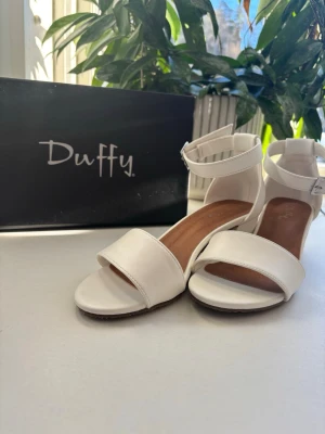 Vita sandaler med klack från Duffy - Stilrena vita sandaler från Duffy med bred rem över tårna och justerbart spänne runt ankeln. Skorna har en låg blockklack och öppen tå, vilket ger en modern och enkel look. Tillverkade i syntetmaterial med mjuk innersula för extra komfort, helt oanvända!