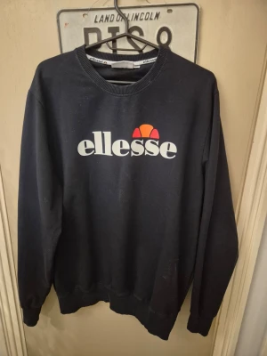 Svart sweatshirt från Ellesse - Säljer en svart sweatshirt från Ellesse med stor vit logga och orange detalj framtill. Klassisk rund halsringning och långa ärmar. Perfekt för dig som gillar sportig och avslappnad stil.