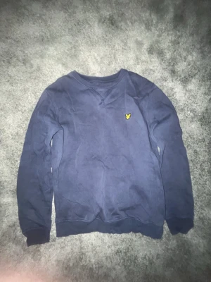 Blå sweatshirt från Lyle & Scott - Mörkblå sweatshirt från Lyle & Scott med klassisk gul logga på bröstet. Tröjan har rund halsringning, ribbade muddar och är långärmad. Perfekt för dig som gillar enkel och stilren design.