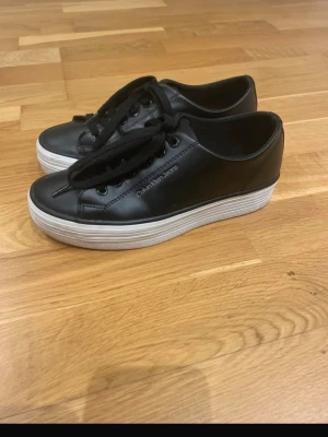 Svarta sneakers från Calvin Klein Jeans - Snygga svarta sneakers från Calvin Klein Jeans med vit, tjock sula och svarta snören. Skorna är i slätt skinnliknande material och har en stilren design med logga på sidan. Perfekta till jeans eller kjol för en clean och modern look. Skorna är använda fåtals gånger och är i väldigt fint skick