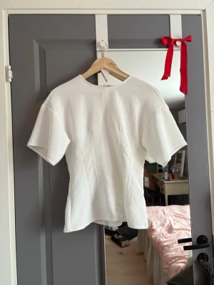 Vit topp med dragkedja från Zara - Säljer en vit topp från Zara i storlek S. Toppen har korta ärmar, rund hals och en synlig dragkedja i ryggen. Materialet är strukturerat och känns som bomull. Snygg passform med markerade sömmar framtill som ger en stilren look. Nästan aldrig använd☺️