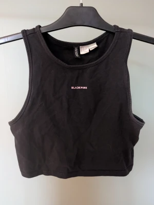 Svart croppad linnetopp BLACK PINK H&M - Säljer en svart croppad linnetopp från H&M med trycket BLACKPINK i rosa framtill. Toppen är ärmlös och har en sportig, enkel design med rund halsringning. Perfekt för dig som gillar K-pop och vill ha en stilren topp till sommaren.