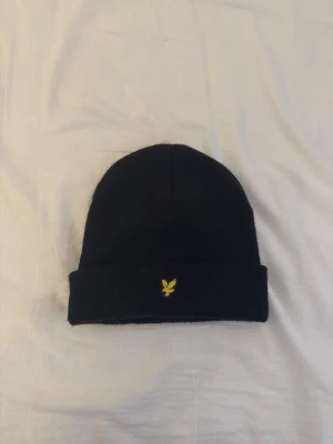 Svart mössa från Lyle & Scott Junior - Säljer en svart mössa från Lyle & Scott Junior med klassisk gul logga framtill. Mössan är ribbstickad och tillverkad i 100% akryl, perfekt för kyliga dagar. Enkel och stilren design som passar till det mesta.