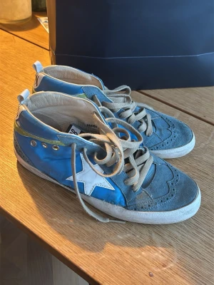 Blå Golden Goose sneakers - Säljer ett par unika sneakers från Golden Goose i blått och grått med vit stjärna på sidan och grönt band bak. Skorna har beige snörning, håldetaljer och GGDB-logga på plösen. Har äkthetsbevis från sellpy.