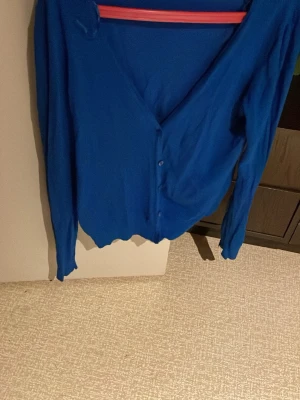 Blå kofta från Lindex - Snygg blå kofta från Lindex med v-ringning och knappar framtill. Långärmad modell i mjukt material, perfekt att slänga över en t-shirt eller linne. Ribbad kant nedtill och vid ärmslut för en clean look. Säljs på fler sidor