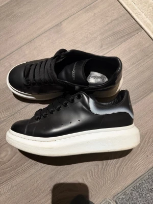 Alexander McQueen svarta sneakers - Säljer ett par feta svarta Alexander McQueen sneakers med chunky vit sula och silvrig detalj vid hälen. Skorna har breda svarta skosnören och är gjorda i skinn. Perfekta om du vill sticka ut med en clean och lyxig vibe.