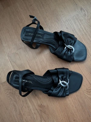 Svarta sandaler med silverdetalj - Säljer ett par svarta sandaler från Maria i storlek 39. De har en fyrkantig tå, låg klack och snygga remmar med en silverring som detalj framtill. Materialet är skinnliknande och sandalerna har en elegant design med knytning runt ankeln.