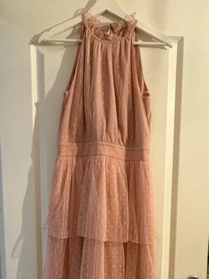 Ljusrosa prickig långklänning med volang - Supersöt ljusrosa långklänning med prickigt mönster och volangdetaljer. Klänningen har ärmlös halterneck-modell med knyt i nacken och dragkedja bak. Tunt, luftigt material som ger en romantisk vibe. Perfekt för sommarens alla tillfällen!