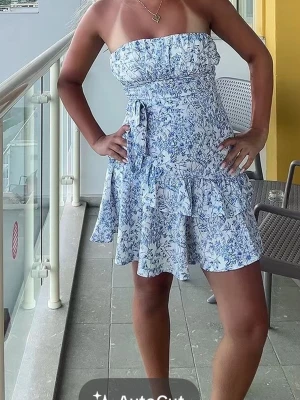 Blommig axelbandslös kortklänning - Söt axelbandslös kortklänning i vitt med blått blommönster. Klänningen har volangkant nertill och ett knytband i midjan. 