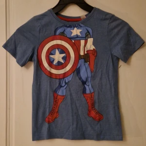 Blå Marvel Captain America t-shirt - Blå t-shirt från Marvel med Captain America-tryck på framsidan. Tröjan har rund hals, korta ärmar och coolt motiv med sköld och röda detaljer. Perfekt för dig som gillar superhjältar. Storlek 122/128. Hund finns i hemmet. Kolla in mina andra annonser för samfrakt! 