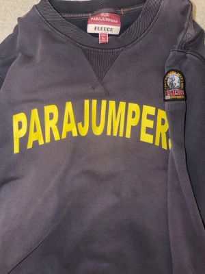 Mörkgrå fleece tröja Parajumpers äkta : storlek L  - Mörkgrå fleece tröja från Parajumpers med stor gul logga över bröstet och patch på ärmen. Ribbstickade muddar och tryck även på insidan av nederkanten. Perfekt för kyliga dagar och riktigt snygg streetstil.