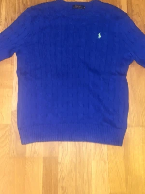 Blå tröja från Polo Ralph Lauren - Säljer en klassisk blå kabelstickad tröja från Polo Ralph Lauren i storlek M. Tröjan har rund hals, lång ärm och den ikoniska polo loggan på bröstet. Perfekt för dig som gillar stilrena och tidlösa plagg.