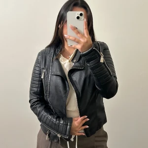 Svart skinnjacka från Voyelles med dragkedjor - Säljer en svart skinnjacka från Voyelles med coola quiltade detaljer på axlar och ärmar. Jackan har flera silvriga dragkedjor, snedställd front och bältesdetaljer vid midjan. Passformen är slim.  Jackan är sliten bakom nacken, på insidan vid ryggen där storleken står, och lite vid ändarna på ärmarna. Skriv för bilder 🥰 