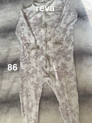 Blommig vit och lila pyjamas - Söt pyjamas i vitt med lila blommigt mönster. Långa ärmar och ben, knäpps framtill med tryckknappar. Perfekt för mysiga dagar hemma eller som pyjamas. 🌸 Fläckar. Reva med hål. Storlek 86