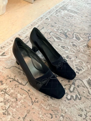 Svarta pumps från Scholl med rosett storlek 38 - Klassiska svarta pumps från Scholl i skinn med mockadetaljer framtill. Skorna har fyrkantig tå, låg klack och en söt rosett på ovandelen. Storlek 38. Använd någon enstaka gång men tyvärr för små efter graviditeten. 