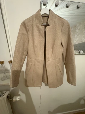 Beige kappa med fiskbensmönster - Snygg beige kappa med diskret fiskbensmönster och stilren krage. Jackan har två fickor framtill och är figursydd för en clean look. Perfekt för dig som gillar klassisk men modern stil. Materialet känns mjukt och har en ullig känsla.