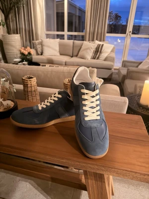 Maison Margiela - Maison Margiela sneakers i marinblått och vitt med beige sula. Skorna har en klassisk rund tå, vita snören och detaljer i både mocka och skinn. Snygg retrostil med kontrasterande material och ikonisk gummisula. Perfekta för dig som gillar stilrena och tidlösa sneakers inför varmare tider. (Obs ej original)