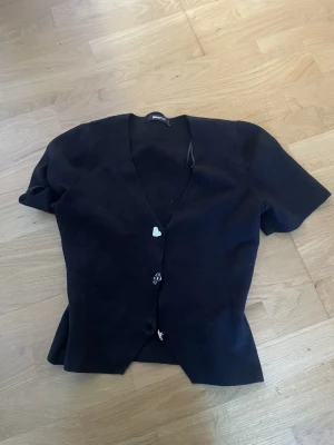 Svart kortärmad kofta från Gina Tricot - Kortärmad svart kofta från Gina Tricot med v-ringning och dekorativa knappar framtill. Tröjan har en enkel och clean look som passar till många olika outfits. Perfekt för lager på lager och snygg till både jeans och kjol.