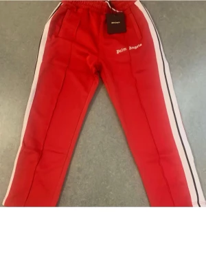 Palm Angels röda mjukisbyxor - Säljer ett par röda mjukisbyxor från Palm Angels med vita och svarta ränder längs sidorna. Byxorna har elastisk midja och fickor framtill. Loggan är broderad på vänster lår. Perfekt för en chill och sportig look.