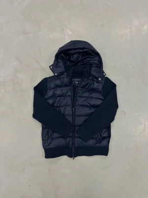 Svart pufferjacka från Leon Marcel - Marinblå cardigan från Leon Marcel med stickade ärmar och huva. Jackan har dragkedja framtill och två sidofickor med dragkedja. Perfekt för dig som gillar en clean och modern look.