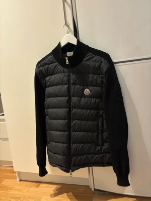 Svart Moncler cardigan jacka L - Säljer en svart cardigan jacka från Moncler i storlek L. Jackan har quiltad front med dunfyllning och stickade ärmar samt sidor. Klassisk Moncler-logga på bröstet och dragkedja framtill. Perfekt för dig som vill ha en stilren och exklusiv look. Kvitto finns köpt från NK! Skulle även kunna tänka mig byte till storlek M