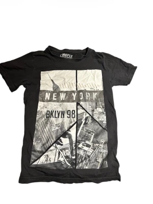 Svart t-shirt Firefly New York print - Svart t-shirt från Firefly med stort grafiskt print på framsidan med texten 'NEW YORK' och 'BKLYN 98'. Trycket har en cool stadssilhuett och skateboardmotiv. Klassisk rund hals och korta ärmar. Tillverkad i mjuk bomull för skön streetwear-vibe. Hör av er vid minsta lilla fundering!😀