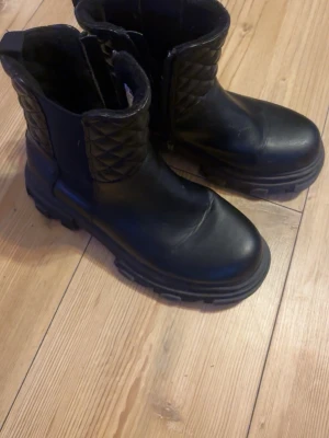 Svarta boots med quiltad detalj - Säljer ett par chunky svarta boots i storlek 36 med quiltad detalj upptill och grov sula. Skorna har dragkedja på sidan för enkel påtagning och är tillverkade i ett skinnliknande material. Perfekta för dig som vill ha en edgy look till hösten eller vintern.