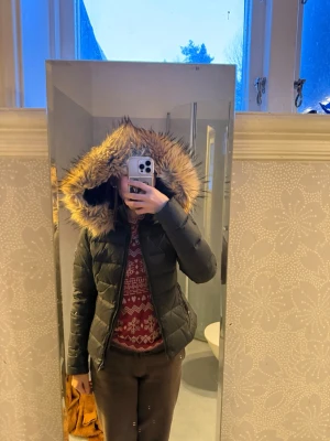 Svart pufferjacka med fuskpäls från Rockandblue - Rockandblue dunjacka köpt 2020 på Design Only. Endast använd två säsonger. Svart, figursydd med huva och fuskpäls. Fint skick, liten lagning finns (se bild). Säljes p.g.a. att den inte används.