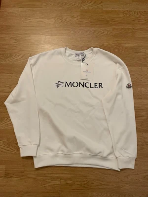 Vit Moncler sweatshirt XL passar L - Säljer en clean vit sweatshirt från Moncler i storlek XL. Tröjan har klassisk rund hals, Moncler-logga broderad på ärmen och stor svart logotyp framtill. Tillverkad i mjuk bomull, perfekt för en stilren och enkel look. Passar L