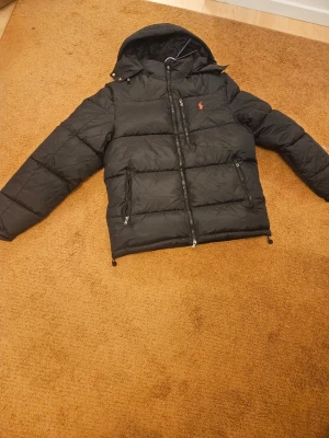 Svart pufferjacka från Polo Ralph Lauren - Säljer en svart pufferjacka från Polo Ralph Lauren med huva och dragkedjor. Jackan har en röd broderad logga på bröstet och flera praktiska fickor med dragkedja. Perfekt för kalla dagar och riktigt snygg streetstil.