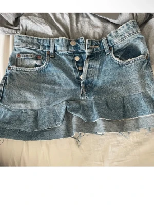 Blå jeanskjol med volang från Zara - Säljer en blå jeanskjol från Zara i storlek 36. Kjolen har volangdetalj nedtill, råa kanter och knappar framtill. Klassisk femficksmodell i denim som ger en avslappnad vibe. Perfekt för dig som gillar en trendig och cool look.