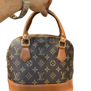  brun handväska  - Säljer en  handväska i klassiskt monogrammönster. Väskan är brun med ljusbruna detaljer och handtag i skinn. Den har guldfärgade metalldetaljer och dragkedja upptill. Perfekt storlek för det viktigaste och en riktig statement piece.