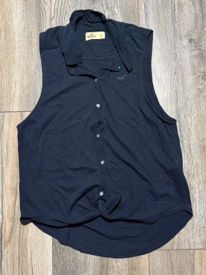 Svart/marinblå ärmlös blus från Hollister - Svart/marinblå ärmlös blus från Hollister med klassisk krage och knappar hela vägen framtill. Toppen har en knytning längst ner för en avslappnad look. Perfekt till jeans eller shorts för en chill stil.